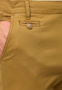 Kakhi hlače s glatkom teksturom, prednjim džepom s kopčanjem na tipku i šavnim detaljima duž ivica.
