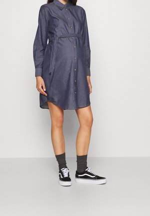 Femme enceinte portant une robe en jean à manches longues boutonnée avec une ceinture nouée à la taille, des chaussettes grises et des baskets noires et blanches, debout à l'intérieur.