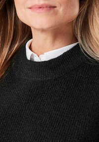 Zwarte textuur trui met een hoge halslijn, gelaagd over een witte blouse met kraag. Close-up die de stofdetails en stiksels toont.