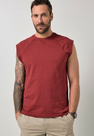 Mann mit Bart, der ein ärmelloses rotes Hemd, beige Hosen, eine Armbanduhr am rechten Handgelenk und einen detaillierten Tattoo-Ärmel am linken Arm trägt.