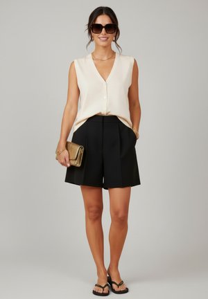 Femme portant un top beige sans manches boutonné, un short noir plissé, des sandales noires à talons, de grandes lunettes de soleil et tenant une pochette dorée.