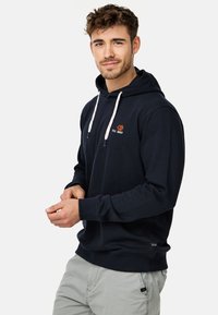 Marineblå hættetrøje med snoretræk, broderet logo på brystet, ribbede manchetter og kant. Blødt materiale, afslappet pasform. Sammen med lysegrå bukser.
