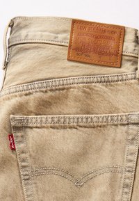 Jean en denim beige Levi's montrant la poche arrière, l'étiquette rouge Levi's et le patch en cuir marron de la marque avec le logo et les détails du texte.