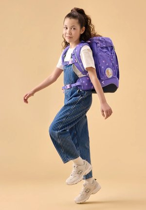 Mädchen in gestreiften Overalls und weißen Turnschuhen, das mit einem großen lila Rucksack mit Punkten und einem Einhorn-Aufnäher vor beigem Hintergrund läuft.