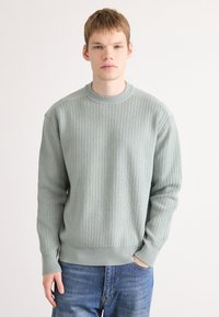 Hellgrüner gestrickter Pullover mit Rippstruktur, Rundhalsausschnitt und langen Ärmeln. Getragen mit blauen Jeans, die sichtbare Nähte zeigen.