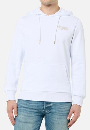 Mann trägt hellblauen Hoodie mit Fronttasche und Kordelzug, kombiniert mit blauen Jeans, vor einem schlichten weißen Hintergrund.