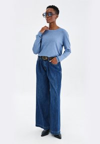 Jasnoniebieska koszula z długim rękawem, luźne niebieskie dżinsowe spodnie z szerokimi nogawkami oraz czarne szpilki z ostrym noskiem. Duże okulary przeciwsłoneczne o unikalnym kształcie.