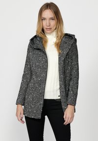 Textúrovaná čierno-biela bunda s kapucňou, s predným zipsom a dlhými rukávmi, v kombinácii s bielym rebrovaným sveterom.