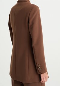 Brun skräddarsydd kavaj med en strukturerad silhuett, skarpa slag och tre knappdetaljer vid ärmarna. Slät tygtextur.