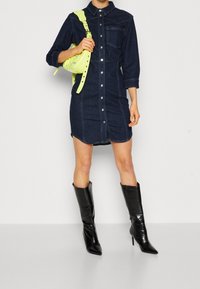 Denim skjortklänning med en figursydd silhuett, mörkblå färg, knäppning fram och långa ärmar. Matcher med en neongul väska och svarta högklackade stövlar.