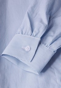 Manica di camicia a strisce sottili blu chiaro e bianche con polsino abbottonato piegata su uno sfondo in tessuto coordinato.