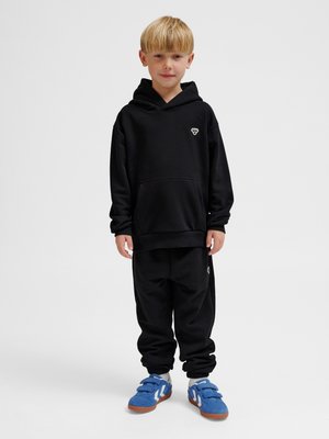 Junger Junge steht, trägt schwarzen Hoodie und Jogginghose mit kleinen weißen Logos sowie blaue Sneakers mit weißen Streifen und braunen Sohlen.