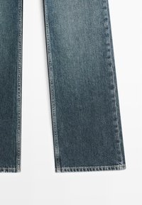 Primo piano delle gambe inferiori di jeans denim blu scuro a taglio dritto con leggera scoloritura e cuciture visibili su sfondo bianco.