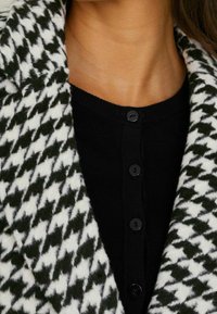 Cardigan en mélange de laine avec un motif pied-de-poule en noir et blanc. Dispose d'un col rond et d'une fermeture à boutons noirs. Texture douce.