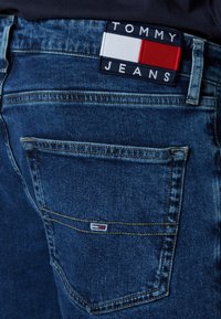 Blåjeans av denim med en mörk tvätt, med detalj på bakfickan och en märkeslapp med röda, vita och blåa färgblock.