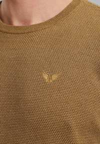 Dzianinowy, brązowy sweter z okrągłym dekoltem. Posiada fakturowany wzór oraz małe złote haftowane logo skrzydła na piersi.