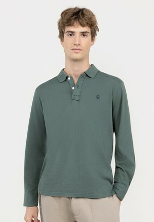 Joven con polo verde de manga larga y pantalones beige, de pie sobre un fondo claro y liso.