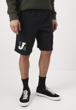 Black athletic shorts feature side pockets and a white and green logo on the left leg. Paired with black sneakers and ribbed black socks.