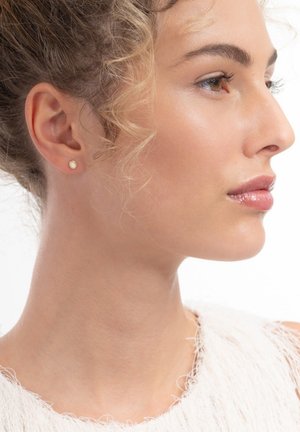 Young woman with curly hair in a white textured top, wearing small round stud earrings, looking to the right against a white background.