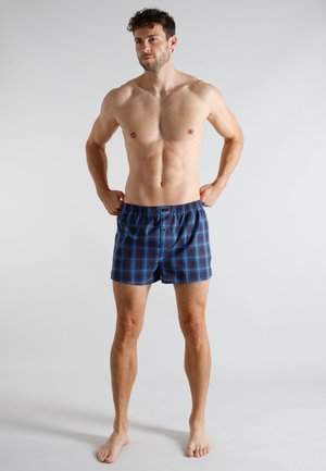 Shirtless man with curly hair wearing blue and black checkered boxer shorts, standing barefoot against plain light background.