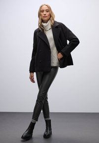 Abrigo negro de doble botonadura, suéter cerrado de punto crema, leggings de cuero sintético negros y botas de tobillo negras con suela gruesa, todo exhibido de manera estilosa.