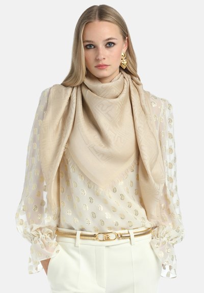 Beige scarf with a subtle pattern, draped elegantly over a white blouse featuring gold polka dots, paired with white pants and a gold belt.