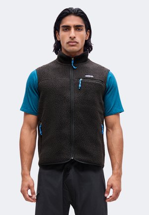 Homme aux cheveux noirs mi-longs porte un gilet polaire noir avec des tirettes de fermeture éclair bleues par-dessus une chemise bleue à manches courtes, debout devant un fond uni.