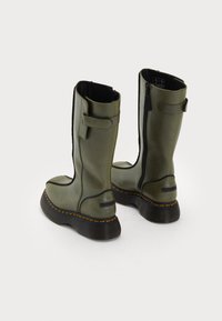 Bottes en cuir vert olive hautes avec des semelles épaisses noires, des coutures orange, des zips sur les côtés et des sangles ajustables sur fond clair.