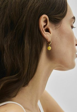 Frau mit langen braunen Haaren, die einen goldenen Creolenohrring mit gelbem Perlenanhänger trägt, seitlich vor einem einfarbigen Hintergrund gesehen.