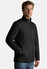 Mężczyzna w czarnej pikowanej kurtce z zamkiem błyskawicznym z przodu, z zamykana kieszenią na piersi oraz dwiema dużymi kieszeniami z przodu, połączonej z niebieskimi dżinsami.