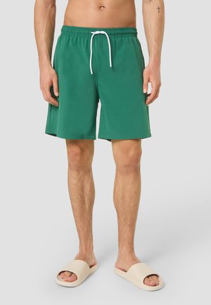 Mand iført grønne badeshorts med hvid snor og beige sandaletter stående foran en ensfarvet baggrund.