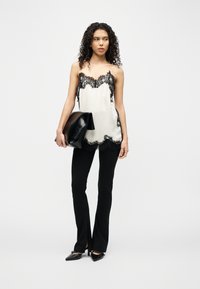 Γυναίκα με σγουρά μαλλιά που φοράει λευκό τοπ με δαντέλα, μαύρο παντελόνι, μαύρα μυτερά ψηλοτάκουνα και κρατάει μια μεγάλη μαύρη τσάντα φάκελο.