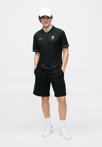 Svart sportsdrakt med vertikale striper, blå aksenter og logo; kombinert med svarte shorts som har mønster av gjentakende tekst; hvite joggesko.