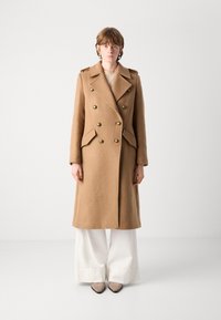 Palton dublu încheiat dintr-un amestec de lână maro, cu butoane aurii, rever mari și buzunare laterale, asortat cu pantaloni albi largi și tocuri taupe.