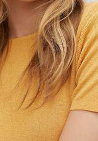 Haut en maille à manches courtes de couleur jaune vif, doté d'un tissu texturé avec une finition douce. La vue de près montre des cheveux qui tombent à proximité.