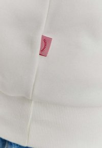 Biela mikina s rebrovaným lemem s malou ružovou nálepkou so jednoduchým dizajnom oka na boku, vyrobená z mäkkej bavlnenej látky.