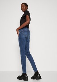 Jeans rectos de color azul oscuro con bolsillos traseros y detalles de costura clara, combinados con zapatos negros y una camiseta negra.