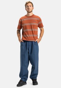 Červeno-oranžové pruhované tričko s krátkým rukávem, v kombinaci s volnými tmavě modrými kalhotami. Černé tenisky dokončují outfit.