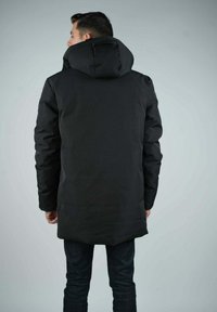 Parka noir isolée avec une grande capuche, des manches longues et une coupe droite, portée par-dessus un jean en denim foncé. Texture lisse et mate.