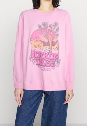 Sudadera rosa con un diseño gráfico que presenta un árbol y una escena desértica. Corte holgado y puños y dobladillo acanalados. Combinada con unos jeans de pierna ancha.