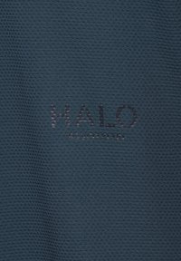 Napis „HALO“ i mniejszy tekst poniżej na ciemnoszarej teksturowanej tkaninie, prawdopodobnie element odzieży sportowej lub casual.