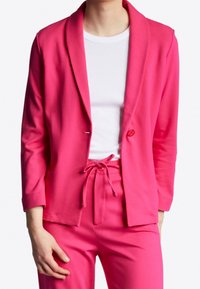 Vaaleanpunainen bleiseri, jossa on shaalikaulus, yksittäinen nappikiinnitys ja pitkät hihat. Yhdistetty samanvärisiin vaaleanpunaisiin housuihin, joissa on nauhavyötärö.