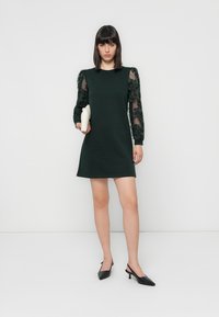 Woman standing in a black dress with sheer, floral-patterned sleeves holding a white clutch, wearing black slingback heels on a white background.