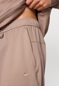 Φόρμες Nike σε ανοιχτό μπεζ χρώμα, με ελαστική μέση, πλαϊνές τσ έπες και μικρό μεταλλικό λογότυπο στο αριστερό μπούτι. Απαλή υφή υφάσματος.