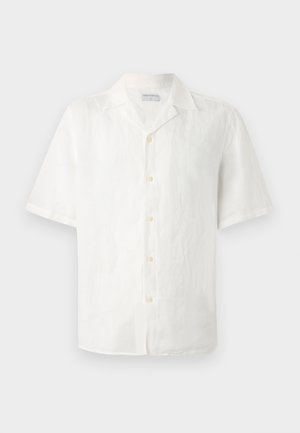 Άνδρας με σγουρά μαλλιά που φοράει λευκό πουκάμισο με κουμπιά και κοντά μανίκια, μπεζ σορτς, λευκές κάλτσες και λευκά αθλητικά, όρθιος με τα χέρια στις τσέπες.