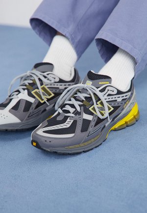 Persona que lleva zapatillas de correr grises, negras y amarillas con calcetines blancos y pantalones azul claro, de pie sobre una superficie azul.