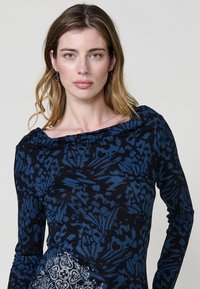 Blusa de manga larga en negro con patrones florales azules, con un escote drapeado y un detalle texturizado en el lado inferior.