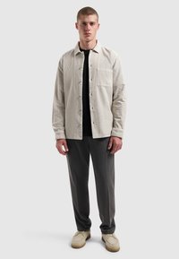 Lichtbeige corduroyshirt met een enkele zak, gedragen over een zwart shirt, gecombineerd met grijze broek en beige schoenen. Eenvoudig, relaxed fit ontwerp.