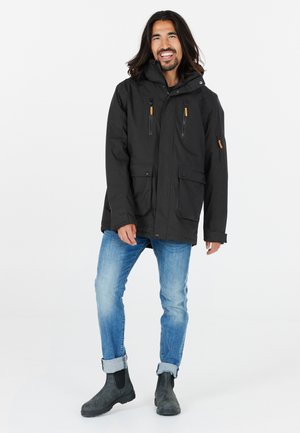 Svart vattentålig jacka med huva, fickor med dragkedja på bröstet och två framfickor med lock. I kombination med blå jeans och grå ankelstövlar.