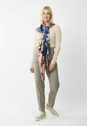 Quadratisches Seidenschal in lebhaften Farben mit abstrakten Mustern, um den Hals gebunden. Helle beige Strickjacke und graue maßgeschneiderte Hose.